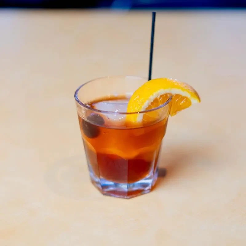Amber cocktail in faceted glass with ice, cherry, and orange slice.