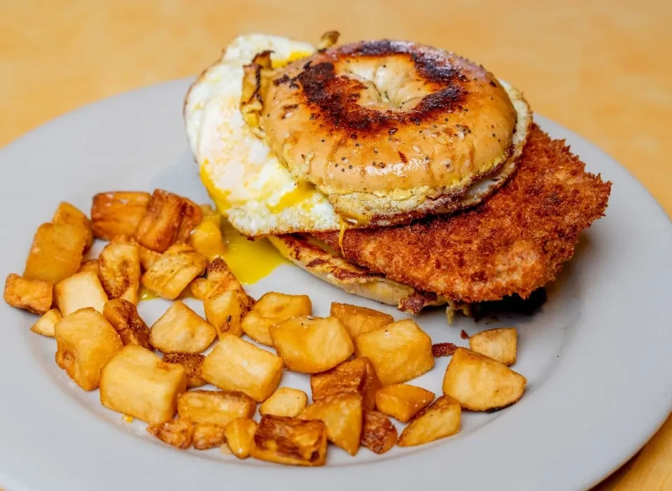 Close-up of a bagel sandwich with egg, schnitzel, and home fries.