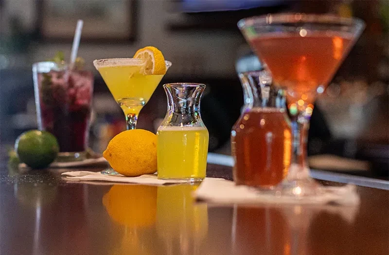 Three colorful cocktails with fruit garnishes on a dark bar counter.