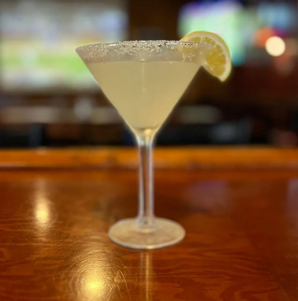 Yellow cocktail in rimmed martini glass with lemon on wooden bar.