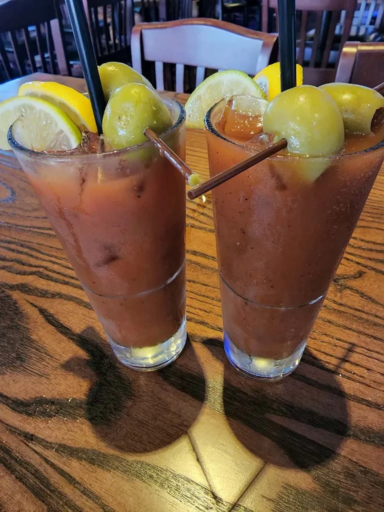 Two reddish-brown cocktails garnished with olives and lemon on a wood table.