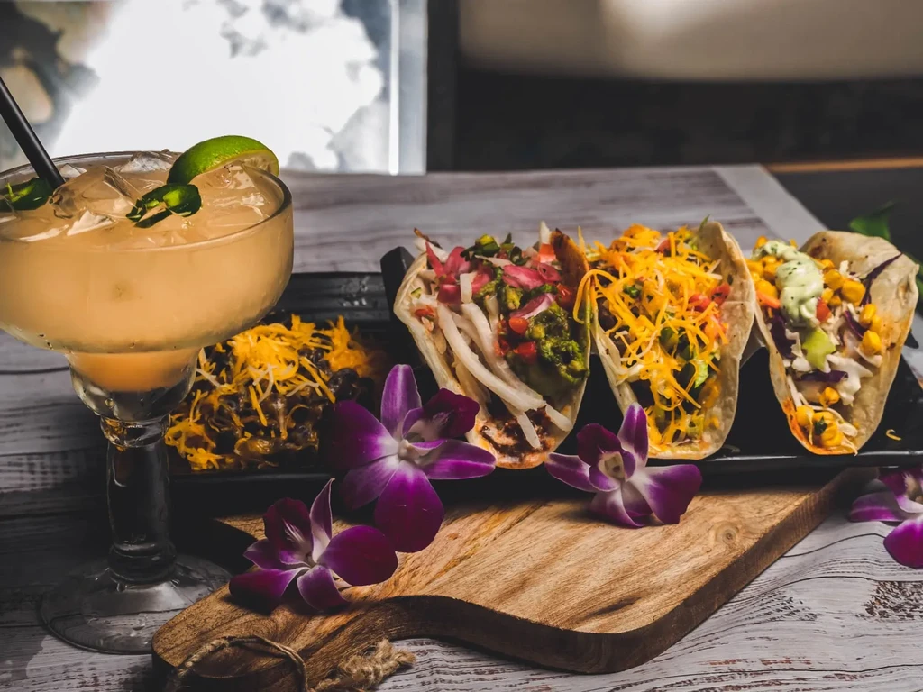 Three colorful tacos, a margarita, and orchids on a wooden table.