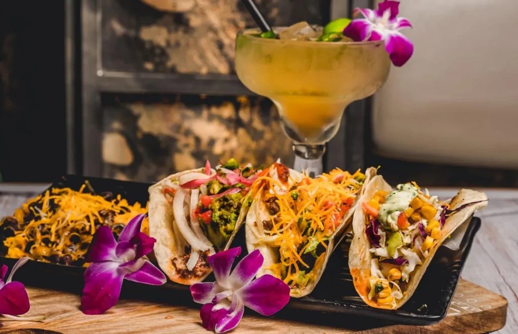Four soft-shell tacos, black beans, and margarita with orchids on wood.