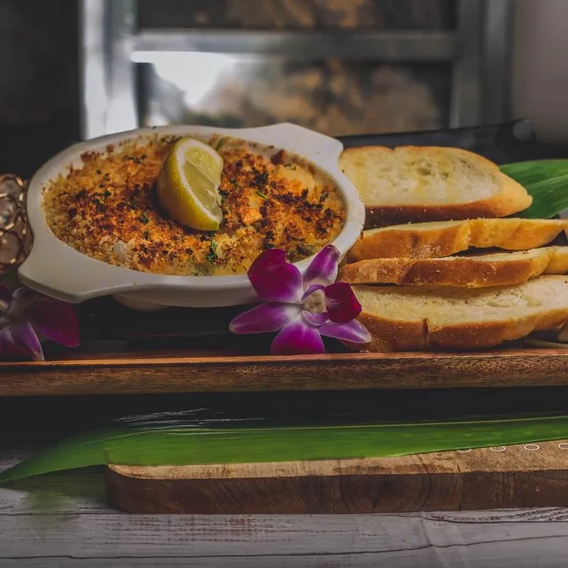 Golden baked dish with lemon, toast, orchids, and tropical leaf.