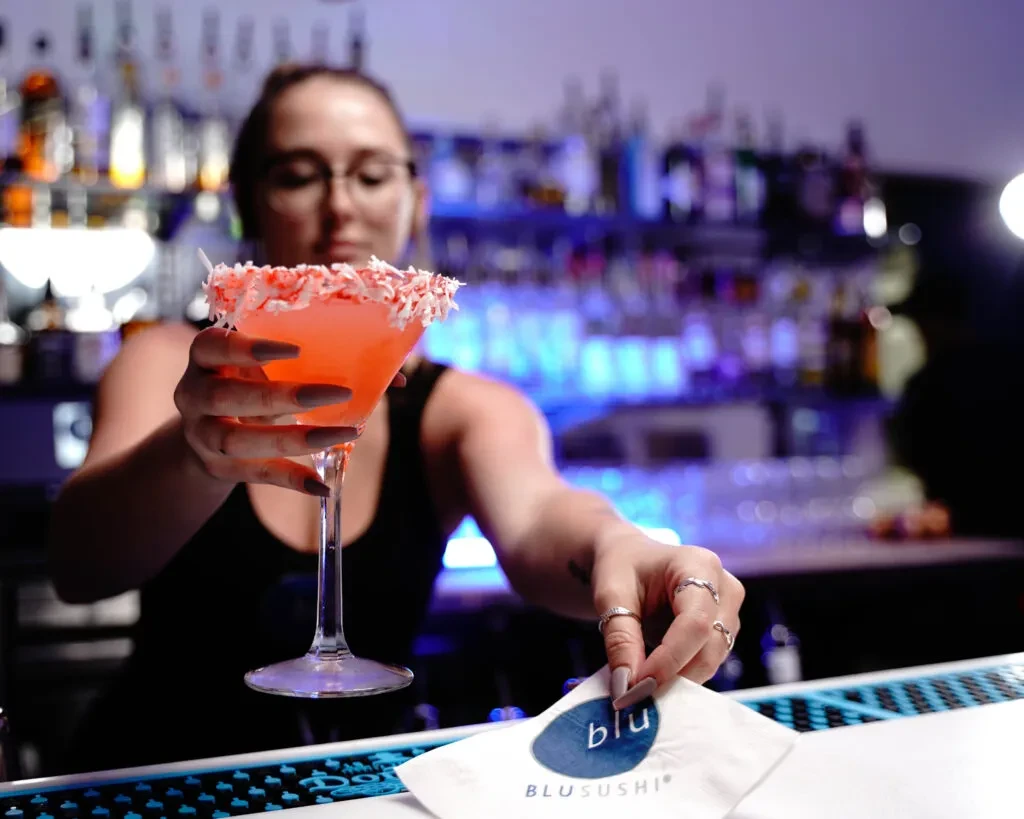 Bartender holds coral-pink cocktail with coconut rim at a modern bar.