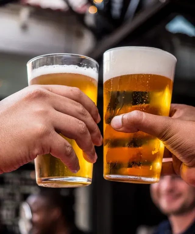 Two hands clink glasses of golden beer with foamy heads.