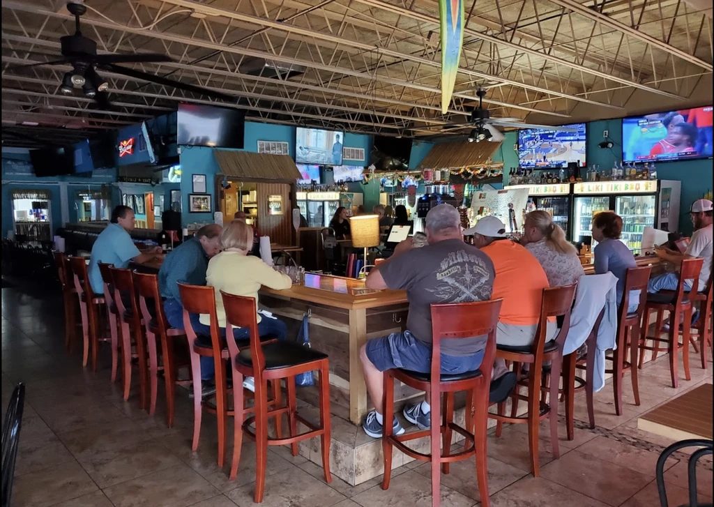Busy tropical bar interior with patrons, screens, and a long counter.