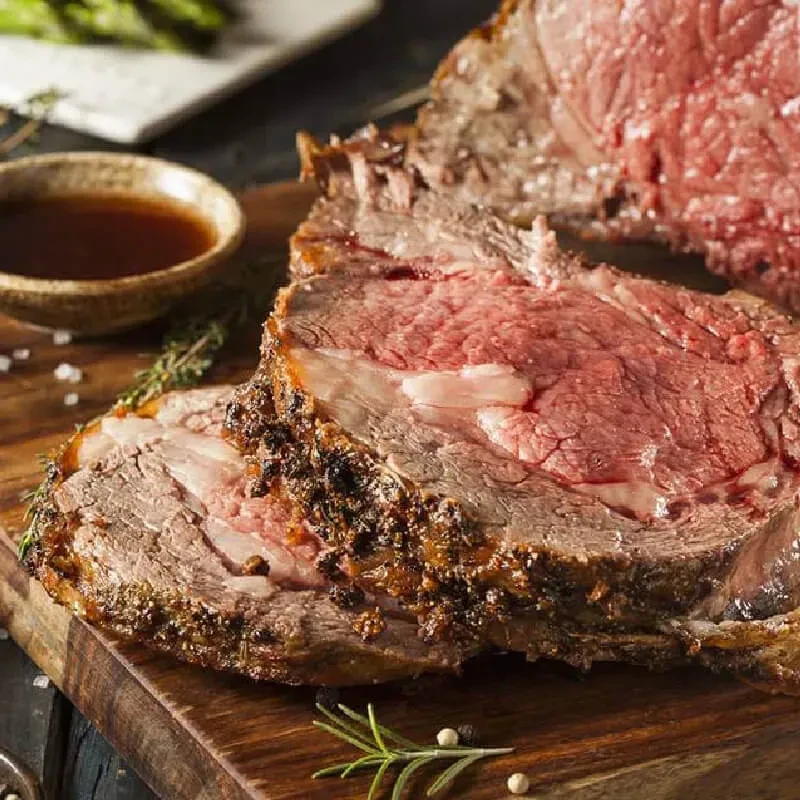 Roasted rare prime rib slice, herbs, and sauce on wooden board.