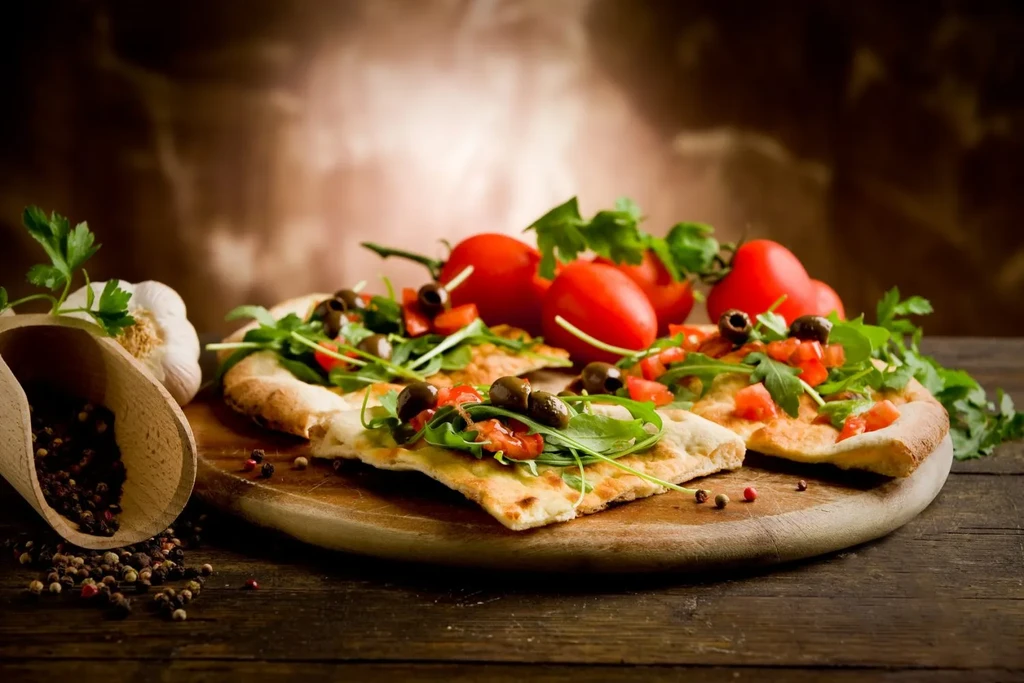 Still life of focaccia with arugula, tomatoes, olives, garlic, and peppercorns.