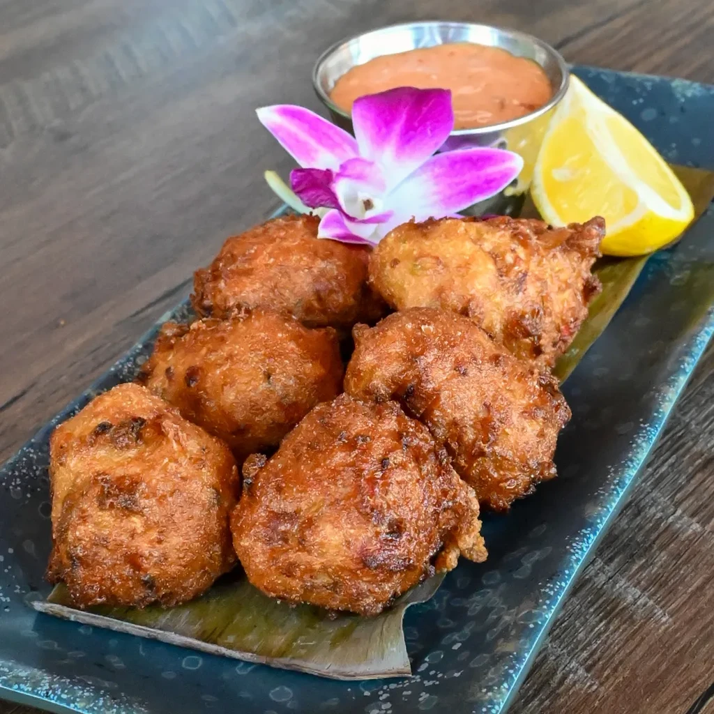 Seven golden-brown fritters on a leaf, with sauce, lemon, and orchid.