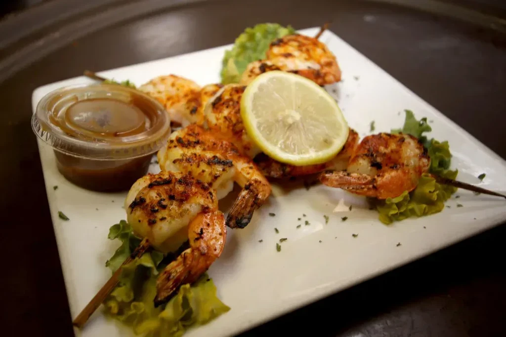 Plate of grilled shrimp skewers with lemon, lettuce, and dipping sauce.