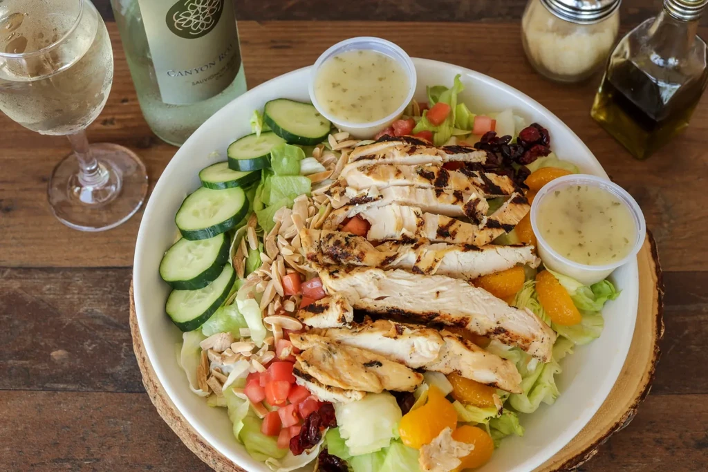 Vibrant chicken salad, dressings, wine, and cheese shaker on a wooden table.