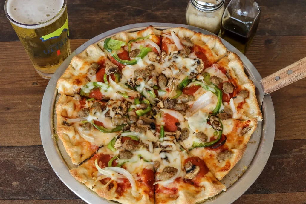 Loaded pizza, cold beer, and condiments on a rustic wooden table.