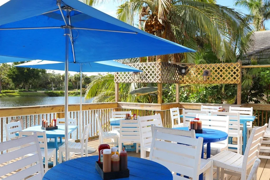 Outdoor restaurant patio: blue tables, white chairs, water, palm trees.