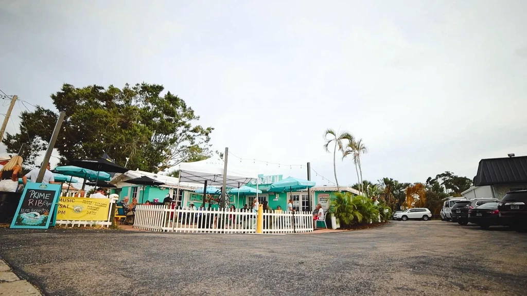 Teal restaurant with outdoor dining patio, people, signs, and palm trees.