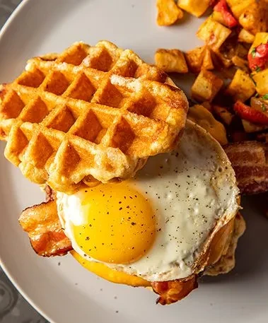 Savory waffle sandwich, fried egg, bacon, and roasted potatoes.