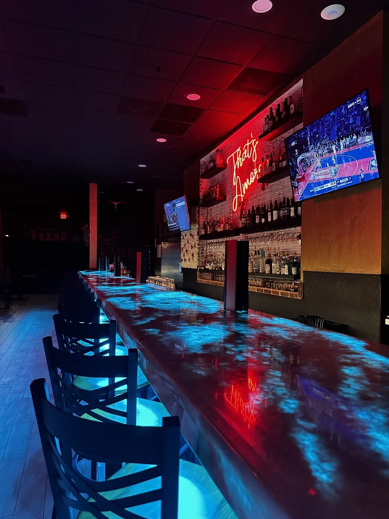 Modern bar interior with glowing blue counter, red neon sign, and TV.