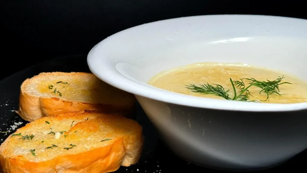 Creamy soup garnished with dill and garlic bread on a dark surface.