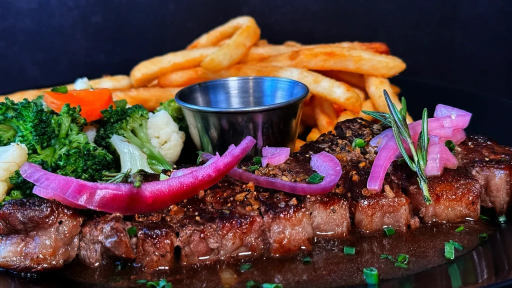 Plated medium-rare steak, sauce, pickled onions, vibrant vegetables, and fries.