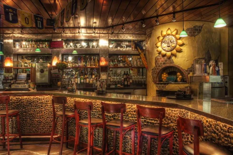 Rustic bar with stone counter, liquor shelves, and brick arch with sun.