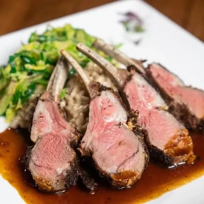 Plated seared medium-rare lamb chops with risotto, greens, and sauce.