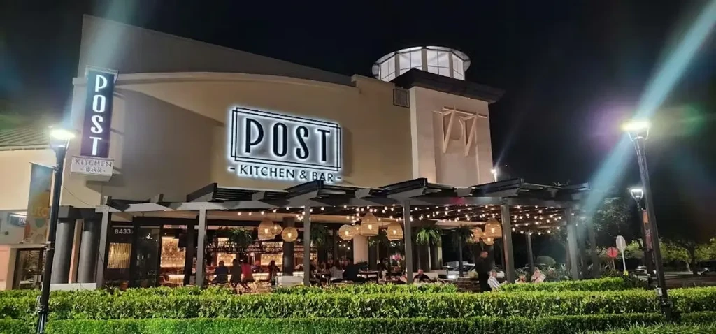 Brightly lit 'POST KITCHEN & BAR' exterior with people dining outside.