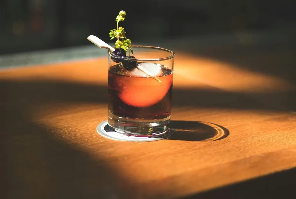 Dramatic cocktail with large ice, berries, herb, and paddle on wood.