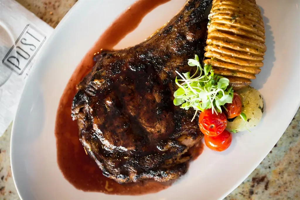 Grilled bone-in steak, Hasselback potato, tomatoes, and microgreens on white plate.