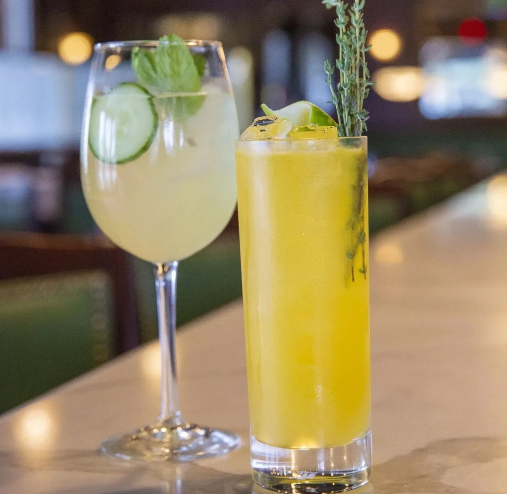 Two cocktails: green with cucumber, yellow with lime, on a light counter.