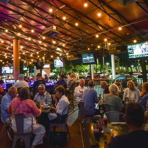 Nighttime bar patio with warm string lights, people, and sports on TVs.