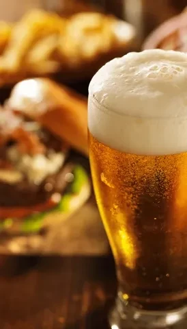 Glass of golden beer with foam, blurry burger and snacks.