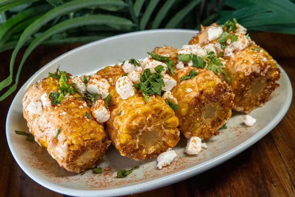 Four grilled corn on the cob with sauce, cheese, herbs on a plate.