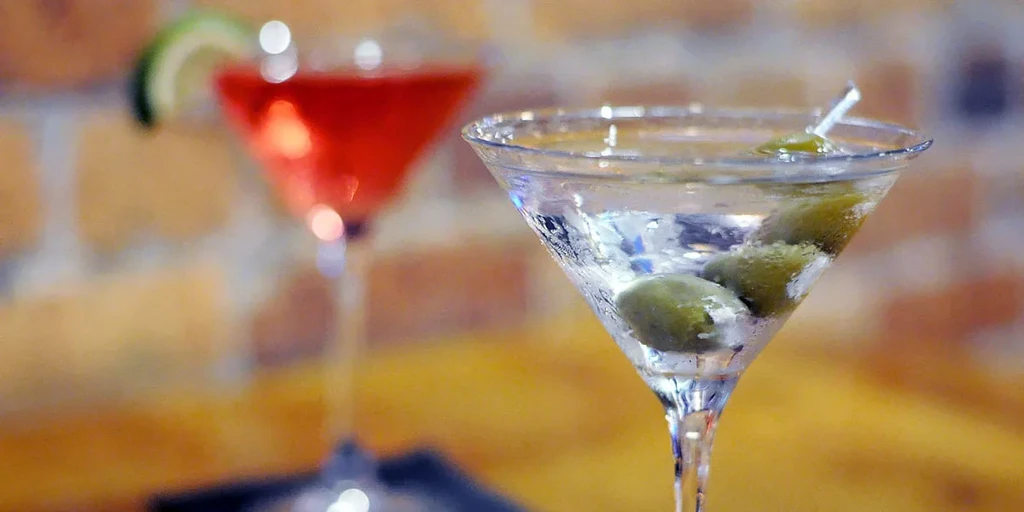 Clear martini with olives, blurred red cocktail with lime behind.