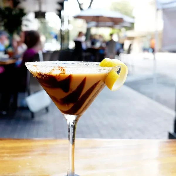 Creamy drink in martini glass, chocolate drizzle, lemon peel, blurred patio.