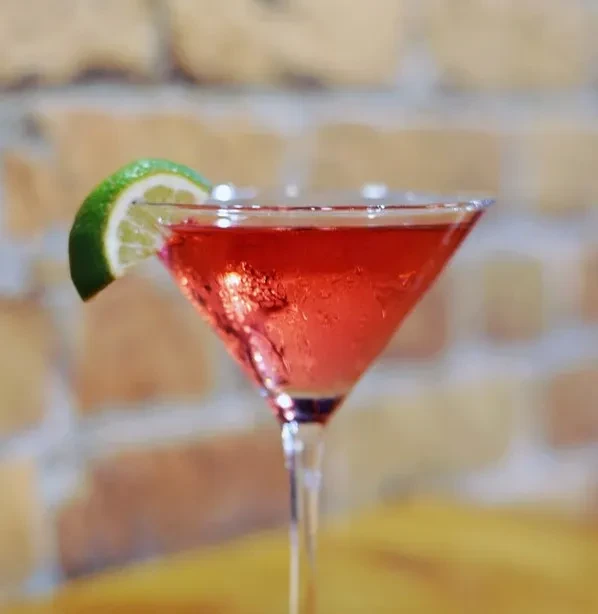 Red cocktail in martini glass with lime wedge, brick wall background.