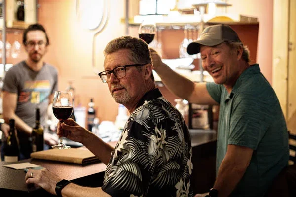 Two men smiling and laughing in a bar with wine.