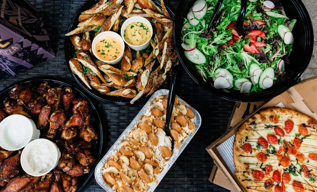 Abundant takeout food spread: pizza, salad, wings, pretzels, dessert.