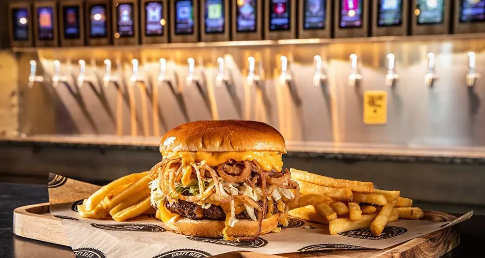 Gourmet burger with fries and onion rings, beer taps in background.