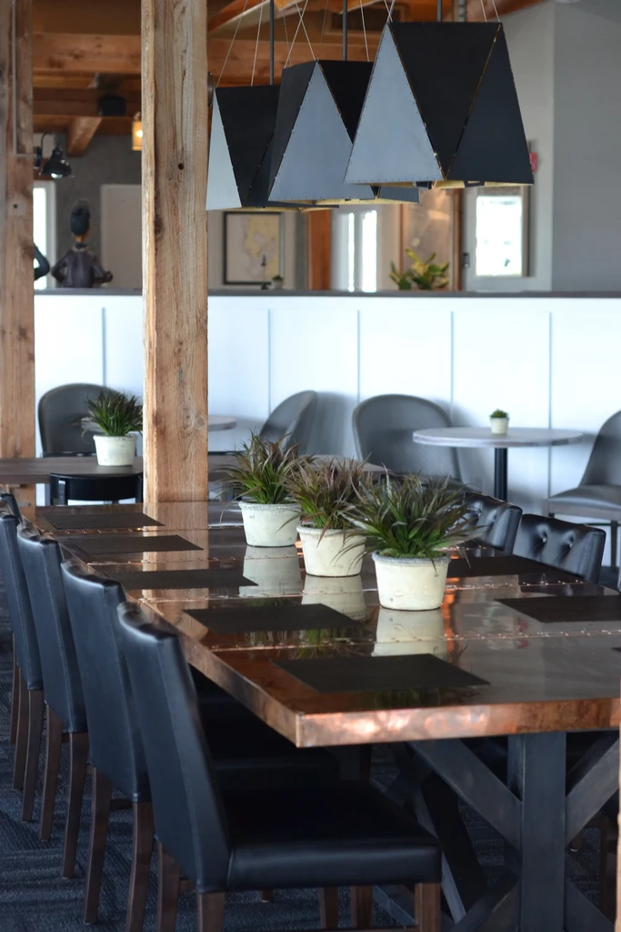 Modern restaurant interior: long reflective table, plants, and geometric lights.