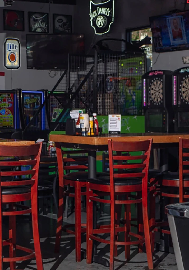 Sports bar interior with arcade games, dartboards, and neon signs.