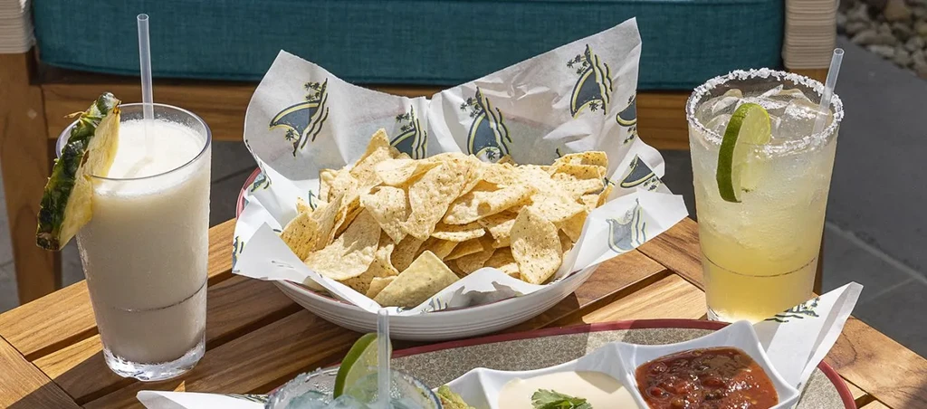 Tortilla chips, guacamole, salsa, and two cocktails on wooden table.
