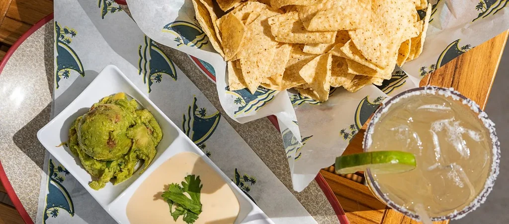 Overhead: tortilla chips, guacamole, creamy dip, and margarita on patterned paper.