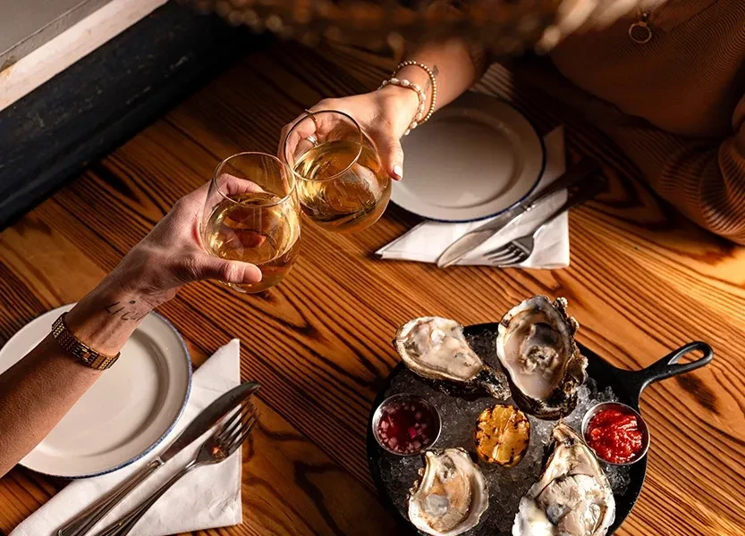 Two hands clink wine glasses over oysters and lemon on a wooden table.