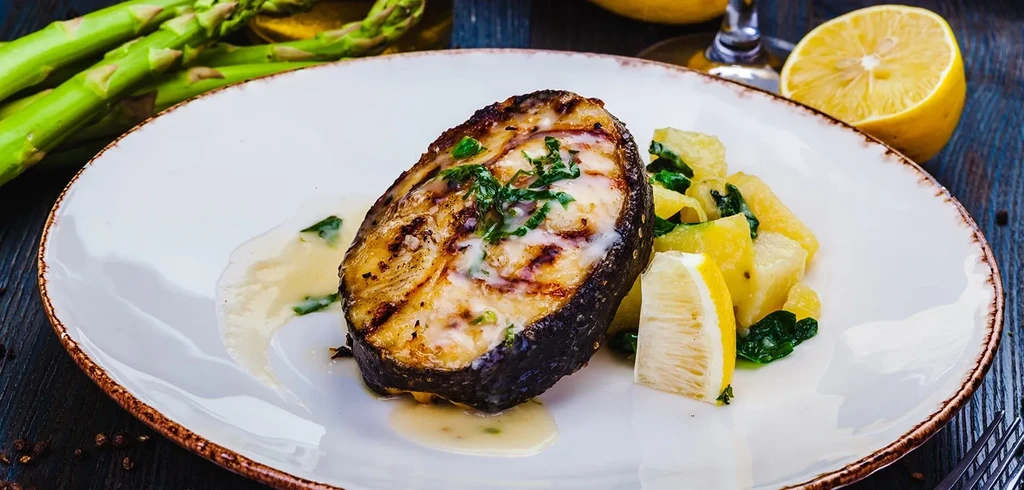 Grilled fish steak with creamy sauce, potatoes, and lemon.