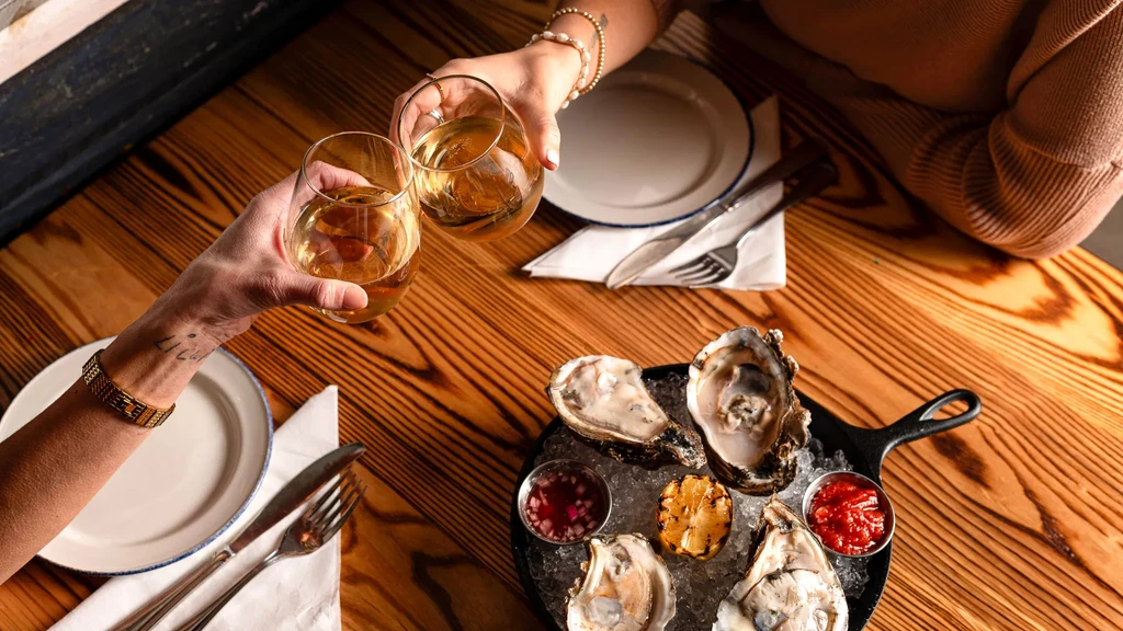 Two hands toast wine above fresh oysters and sauces.