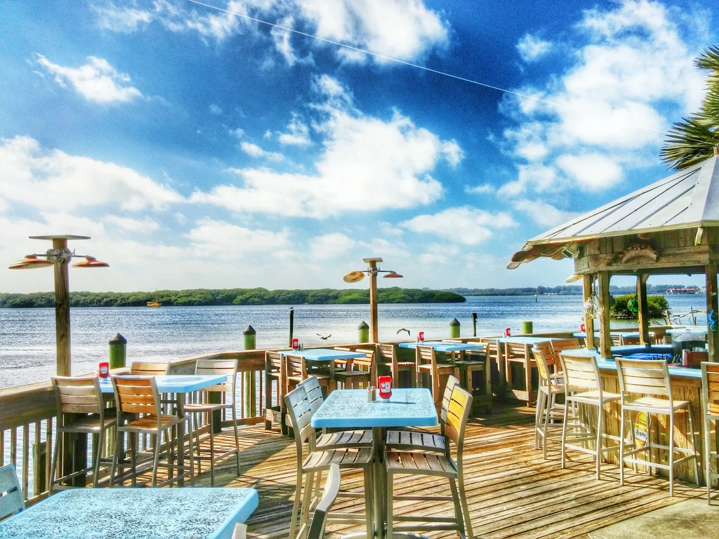 Sunny waterfront restaurant deck, empty tables, bar, distant boats.