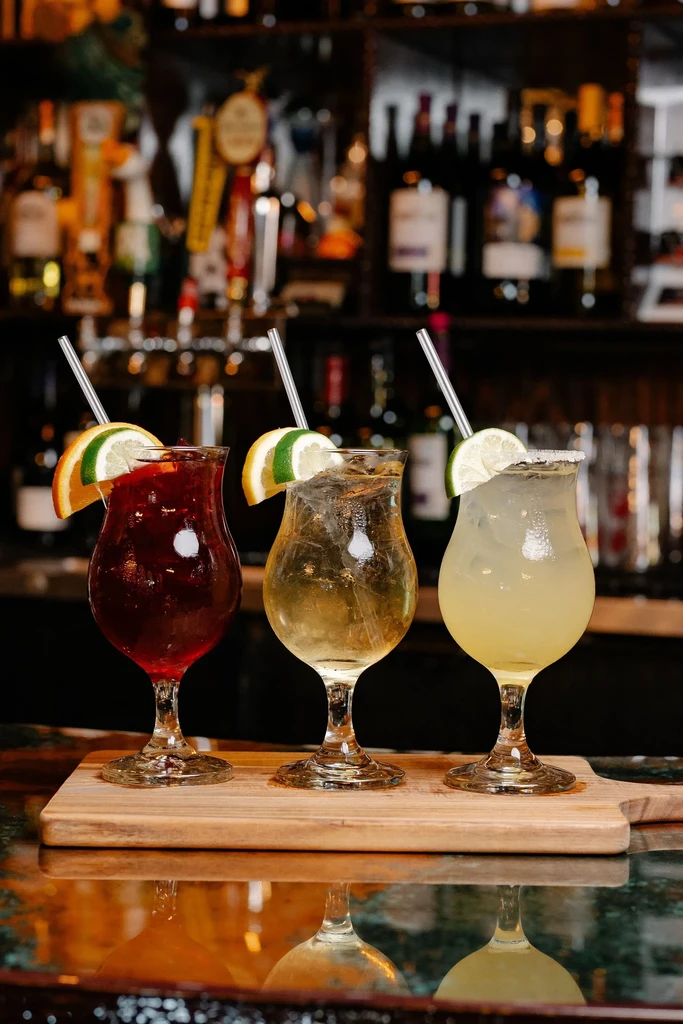 Three colorful garnished cocktails on a wooden board in a bar.