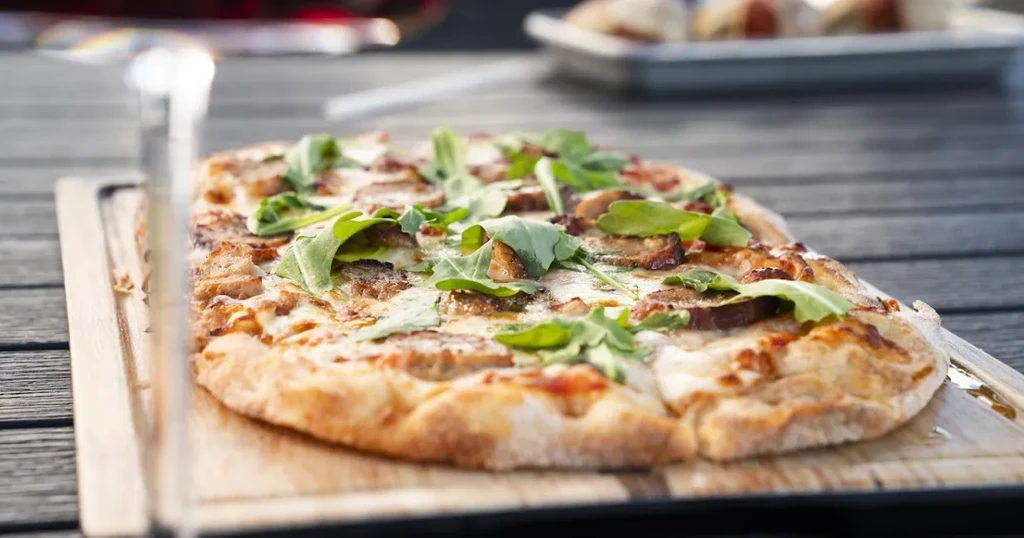 Pre-sliced rectangular flatbread pizza with cheese, meat, and arugula.