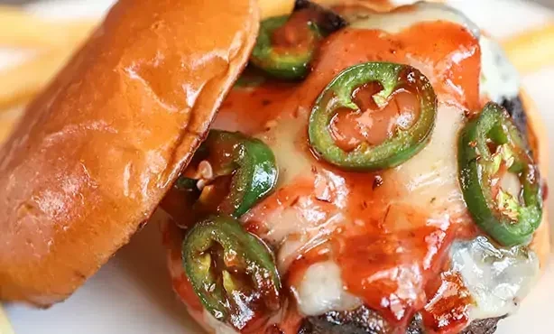 Close-up of a burger with melted cheese, red sauce, and jalapeños.