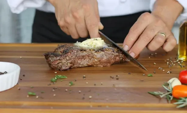 Hands slicing seared steak with melting herbed butter on a wooden board.
